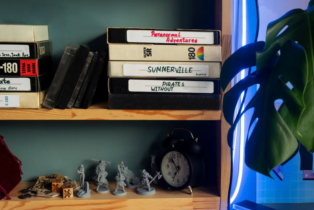 Drevená polička s VHS kazetami, stolnými miniatúrami postáv, budíkom a izbovou rastlinou.